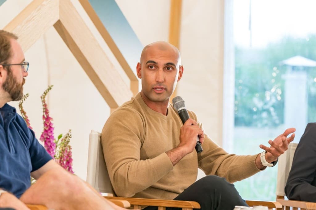 A man holding a microphone speaks while seated on a panel with two other style experts during a discussion event.